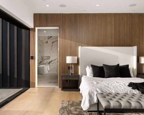 Modern bedroom interior with wooden accent wall, white bed, and open sliding glass door leading to a balcony.